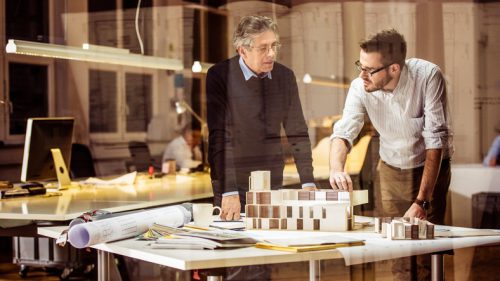Young architect explaining project plan to senior coworker in the office with the help of an architectural model, shot through glass.